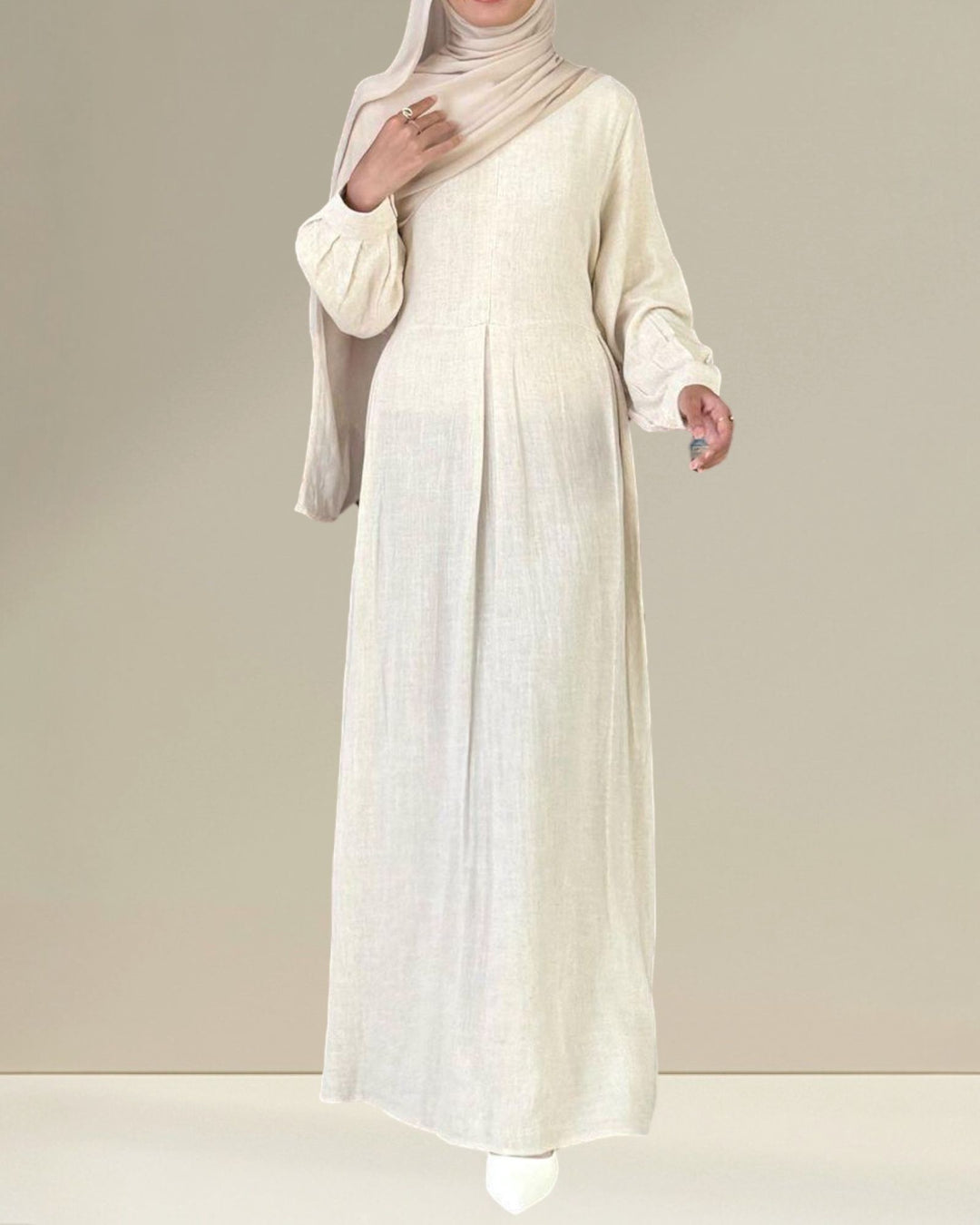 Woman wearing a long, light-colored dress with a matching hijab on a plain background